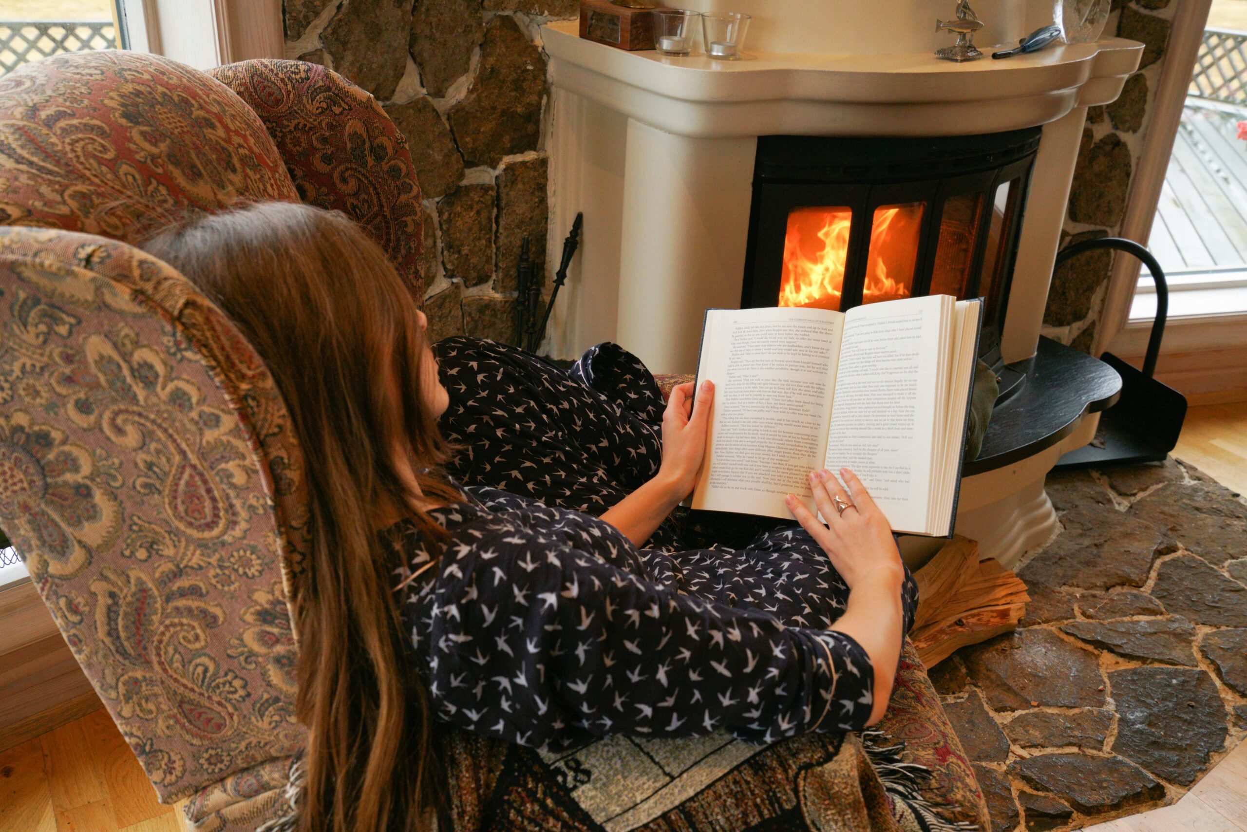 Woman relaxing and reading by a warm fireplace in a comfortable armchair.