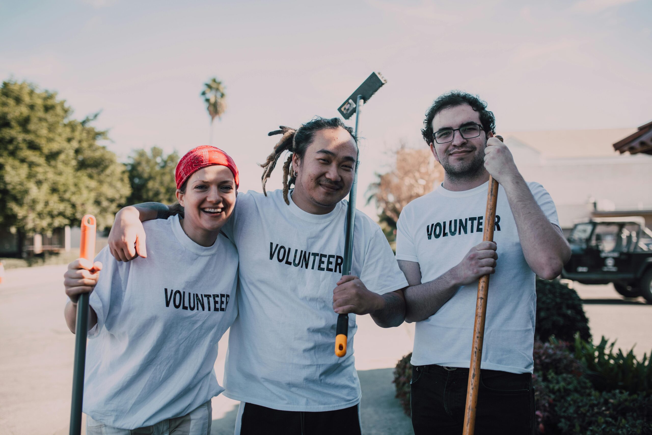 Three diverse volunteers smiling and holding cleaning tools during a community service event.