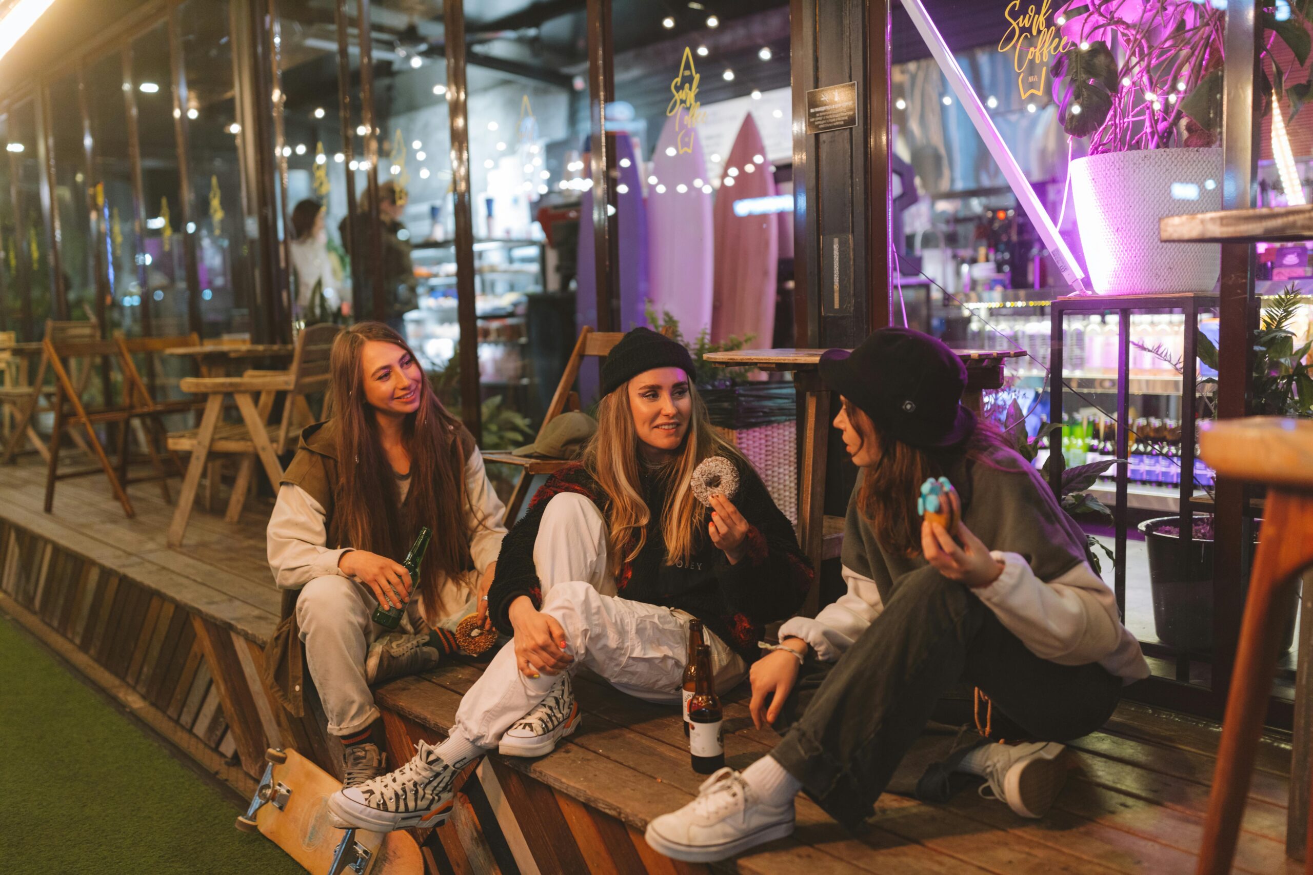 Three young women enjoy donuts and drinks outside a trendy cafe at night, sharing a moment of relaxation and friendship.