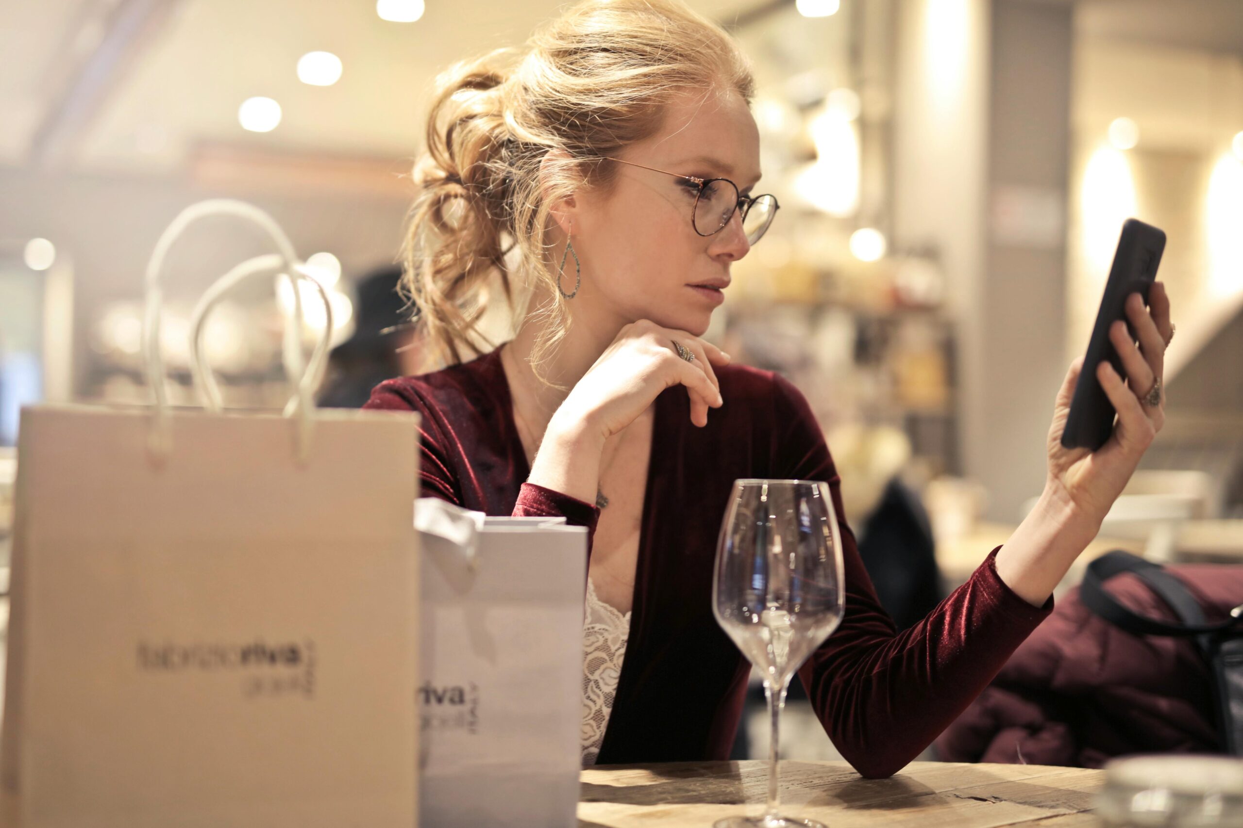 Elegant woman in a restaurant checking her smartphone, surrounded by shopping bags and a wine glass.